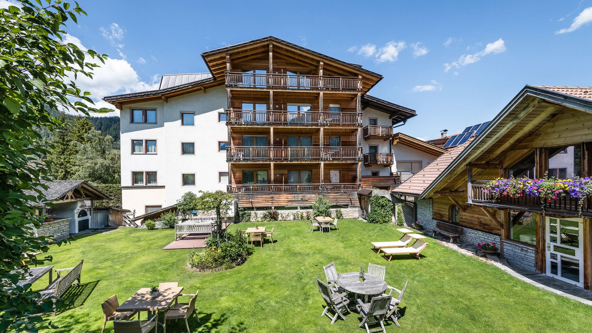 Haus mit Holzbalkonen und Garten mit Tischen und Liegen an sonnigem Tag