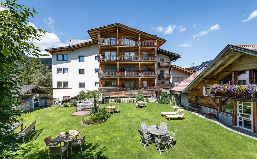 Haus mit Holzbalkonen und Garten mit Tischen und Liegen an sonnigem Tag