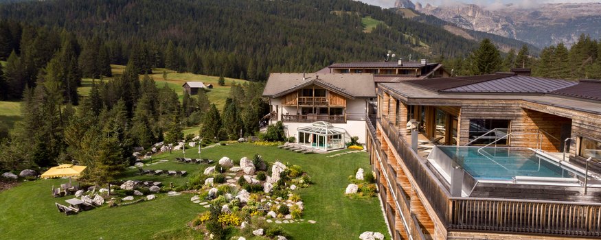 Hotel mit Außenpool auf Terrasse und grüner Wiese vor Bergwald