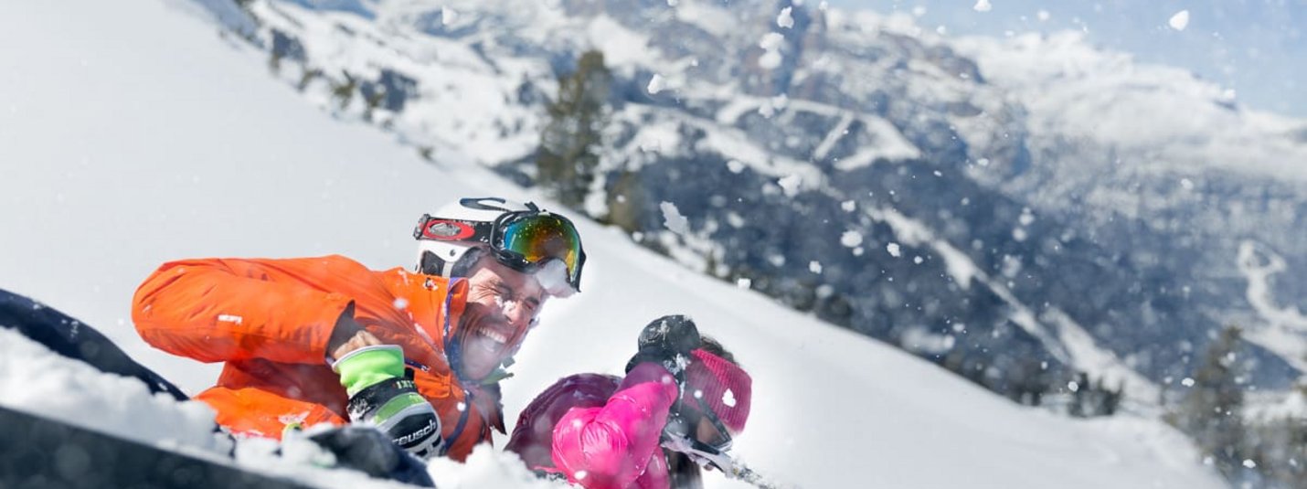 Two skiers fall in snowy mountains throwing snow at each other