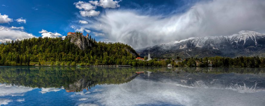 Burg auf bewaldetem Hügel mit See und schneebedeckten Bergen im Hintergrund