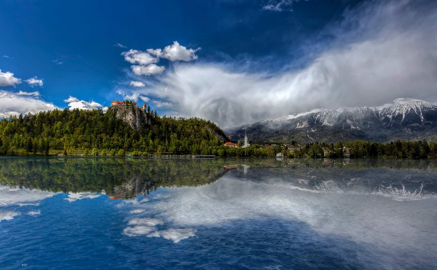 Castello su collina boschiva con lago e montagne innevate sullo sfondo