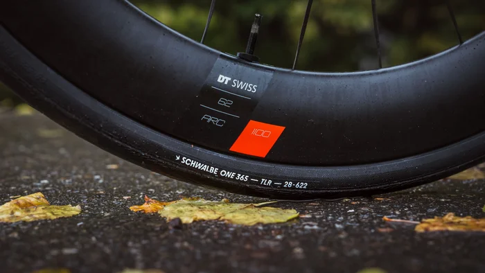 Schwalbe One 365 © Ralf Bohle GmbH Nahaufnahme eines Fahrradreifens auf nassem Asphalt zwischen herbstlichen Blättern