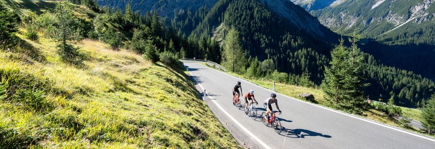 Three cyclists on mountain road surrounded by mountains and green grass