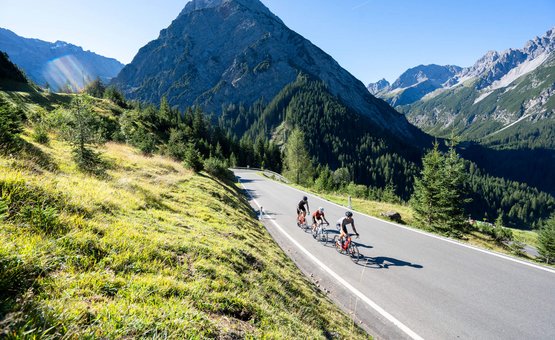 Three cyclists on mountain road surrounded by mountains and green grass