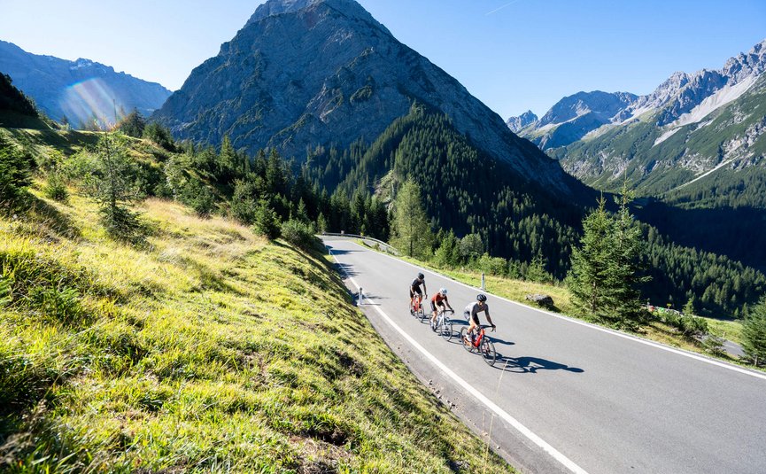 Three cyclists on mountain road surrounded by mountains and green grass