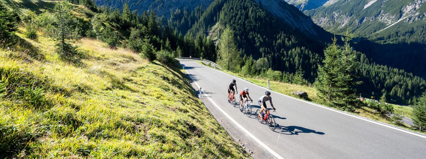 Drei Radfahrer auf Bergstraße umgeben von Bergen und grünem Gras