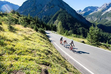 Tre ciclisti su strada di montagna circondati da montagne e erba verde