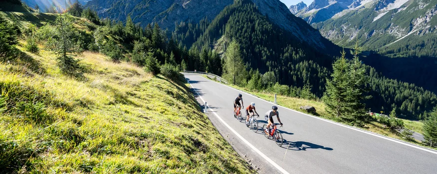 Drei Radfahrer auf Bergstraße umgeben von Bergen und grünem Gras