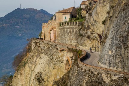 Cyclist on steep road by rocky cliff with mountain and houses in background