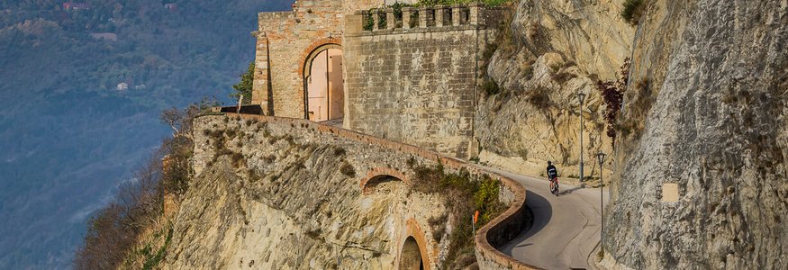 Ciclista su strada ripida vicino a scogliera rocciosa con montagna e case sullo sfondo