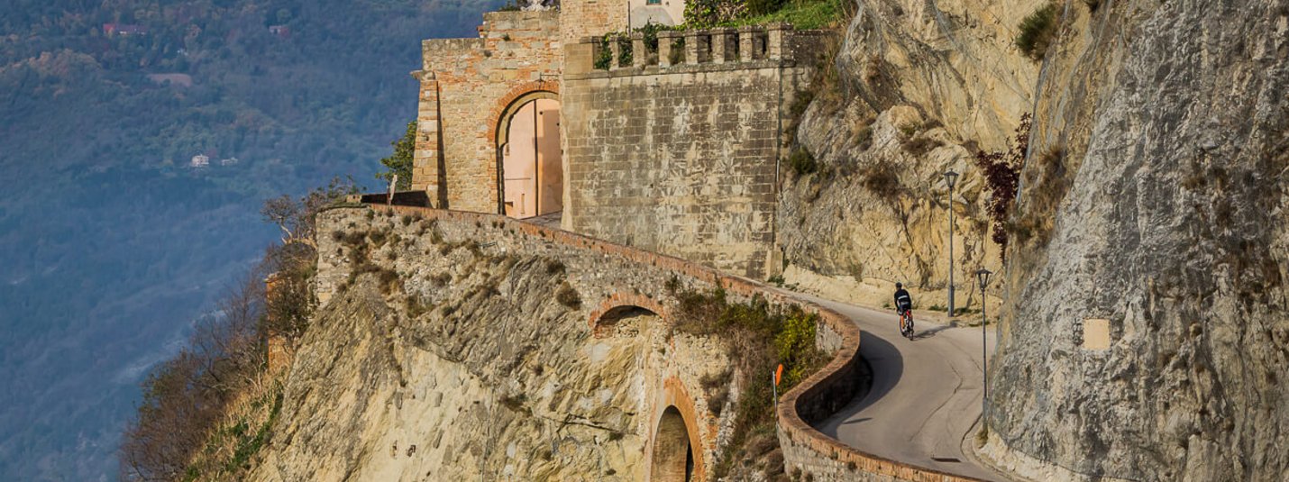 Ciclista su strada ripida vicino a scogliera rocciosa con montagna e case sullo sfondo