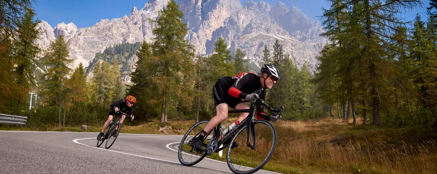 Zwei Radfahrer fahren auf kurviger Bergstraße mit Bäumen und Bergen im Hintergrund