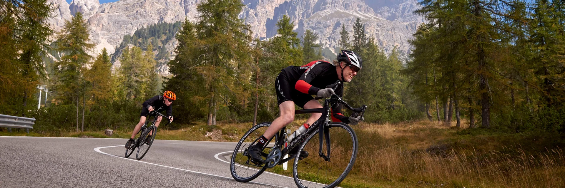 Zwei Radfahrer fahren auf kurviger Bergstraße mit Bäumen und Bergen im Hintergrund