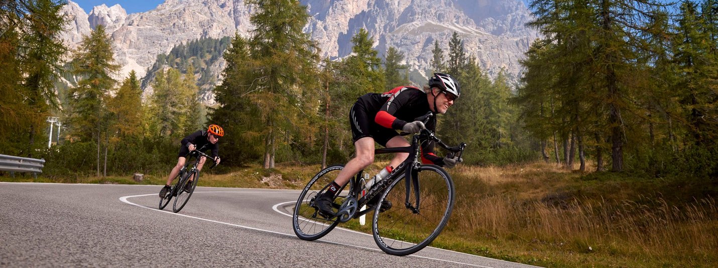 Zwei Radfahrer fahren auf kurviger Bergstraße mit Bäumen und Bergen im Hintergrund