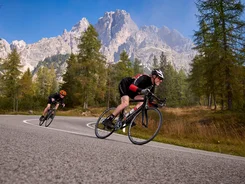 Two cyclists riding on a winding mountain road with trees and mountains behind