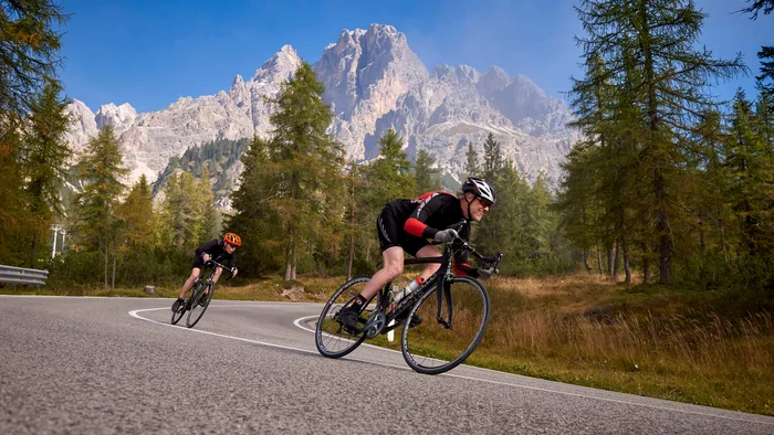 Die schönsten Rennrad-Regionen und Tipps für den Herbst © Joris Lugtigheid Zwei Radfahrer fahren auf kurviger Bergstraße mit Bäumen und Bergen im Hintergrund