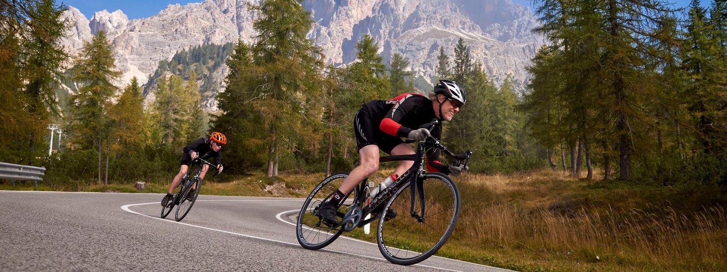 Due ciclisti su strada di montagna tortuosa con alberi e montagne sullo sfondo
