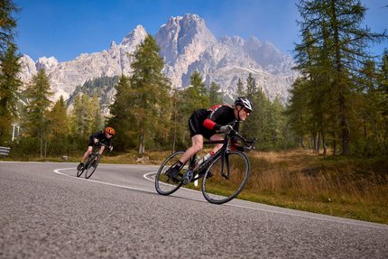 Two cyclists riding on a winding mountain road with trees and mountains behind