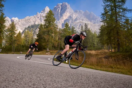 Zwei Radfahrer fahren auf kurviger Bergstraße mit Bäumen und Bergen im Hintergrund