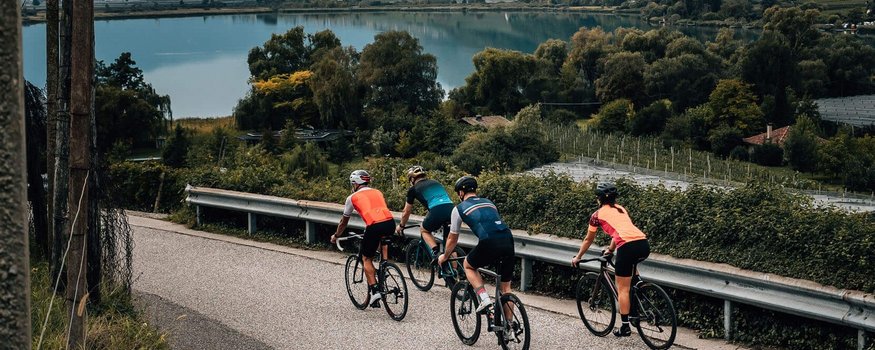 Vier Radfahrer fahren auf einer Straße neben einem See und Bergen