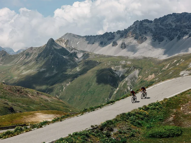 Zwei Radfahrer fahren einen Bergpass mit Alpen im Hintergrund hinauf
