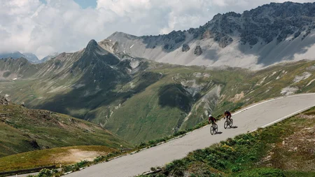 Zwei Radfahrer fahren einen Bergpass mit Alpen im Hintergrund hinauf
