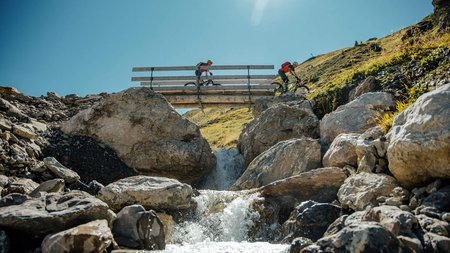 Zwei Mountainbiker fahren über eine Brücke über einen Bach in den Bergen