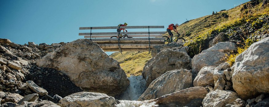 Auf Schmugglerpfaden durch Ischgl/Samnaun
© David Karg Zwei Mountainbiker fahren über eine Brücke über einen Bach in den Bergen