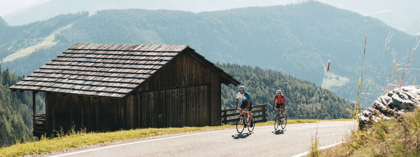 Two cyclists riding on mountain road next to wooden hut in the Alps