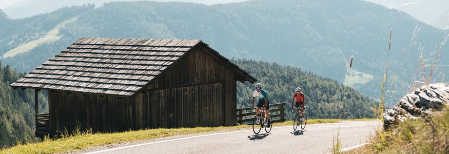 Two cyclists riding on mountain road next to wooden hut in the Alps