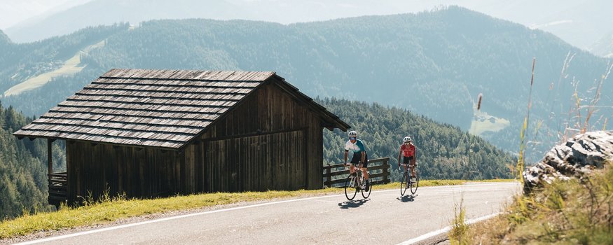 Two cyclists riding on mountain road next to wooden hut in the Alps