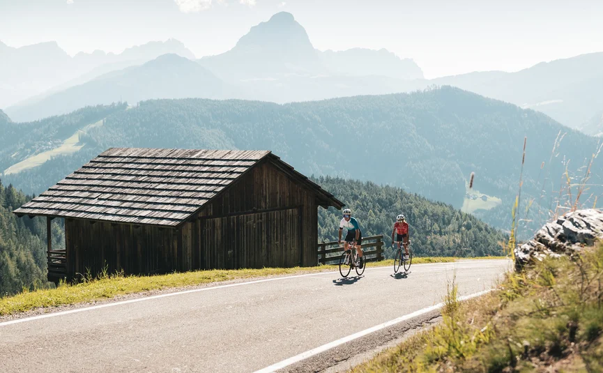Two cyclists riding on mountain road next to wooden hut in the Alps