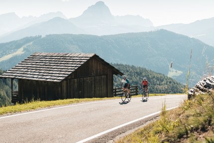 Twee wielrenners op bergweg naast houten hut in de Alpen