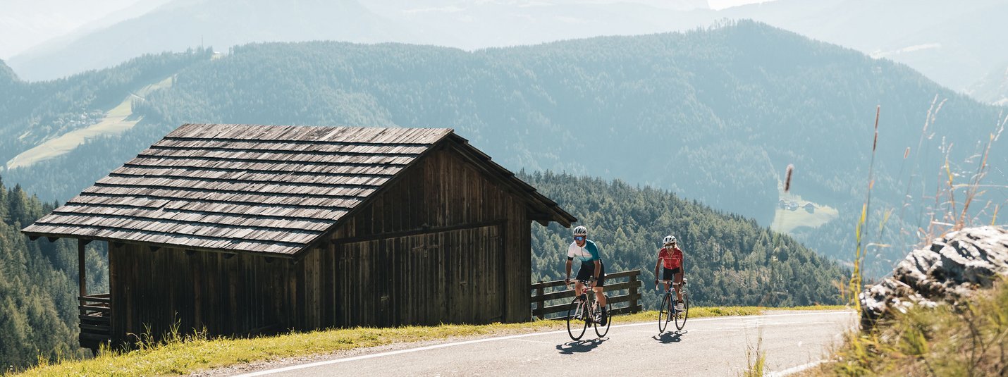 Zwei Radfahrer fahren auf Bergstraße neben Holzhütte in den Alpen