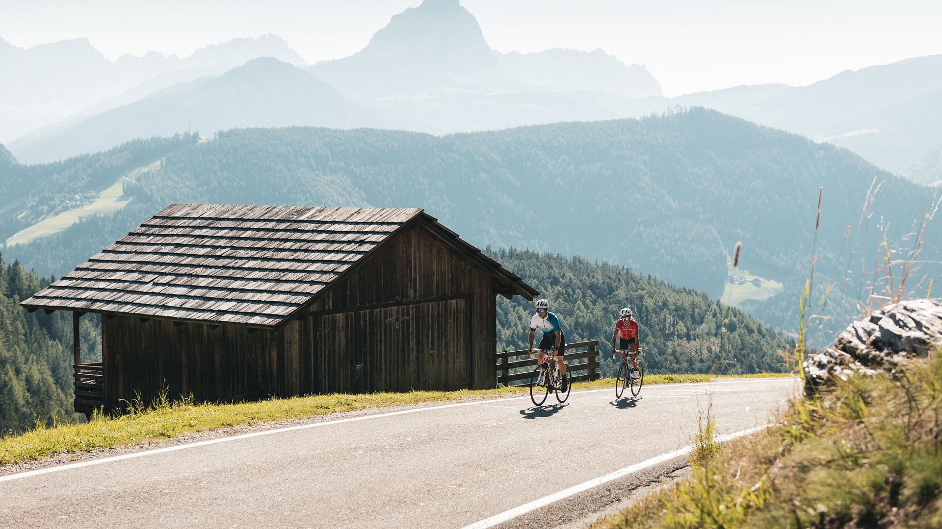 Zwei Radfahrer fahren auf Bergstraße neben Holzhütte in den Alpen