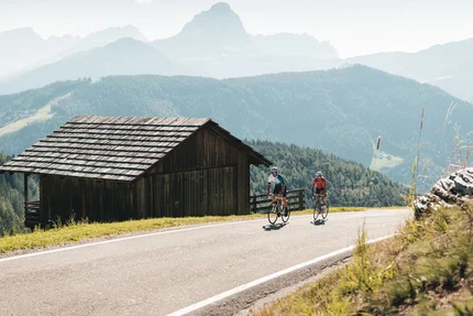Zwei Radfahrer fahren auf Bergstraße neben Holzhütte in den Alpen