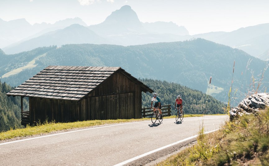 Two cyclists riding on mountain road next to wooden hut in the Alps