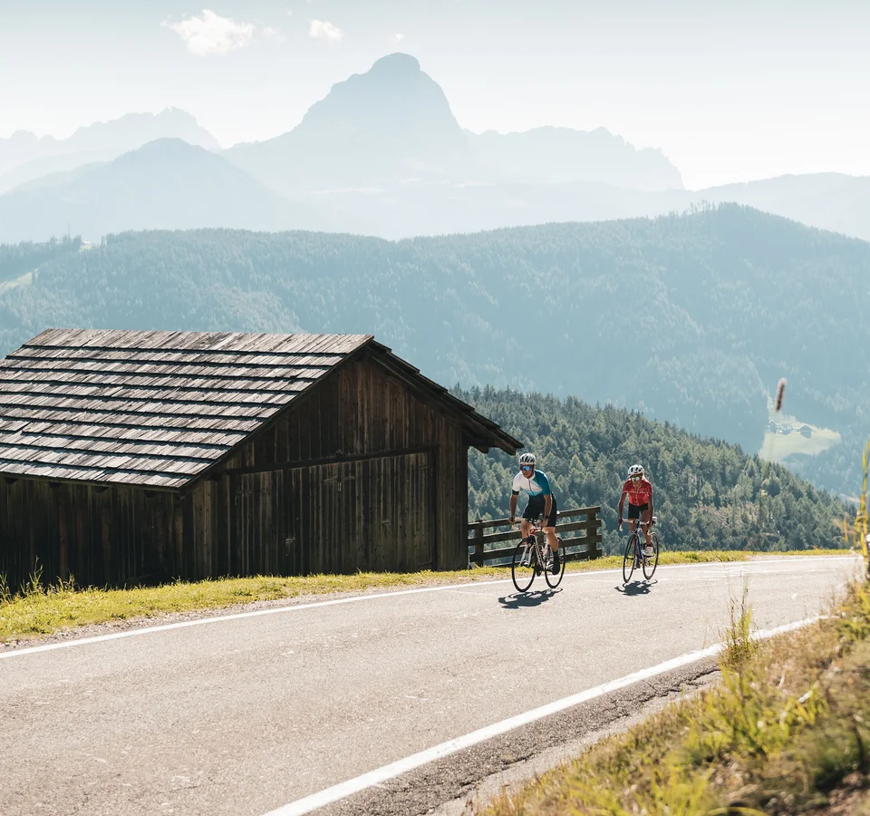 Kronplatz: Höhenmeter ohne Ende © Alex Moling Zwei Radfahrer fahren auf Bergstraße neben Holzhütte in den Alpen
