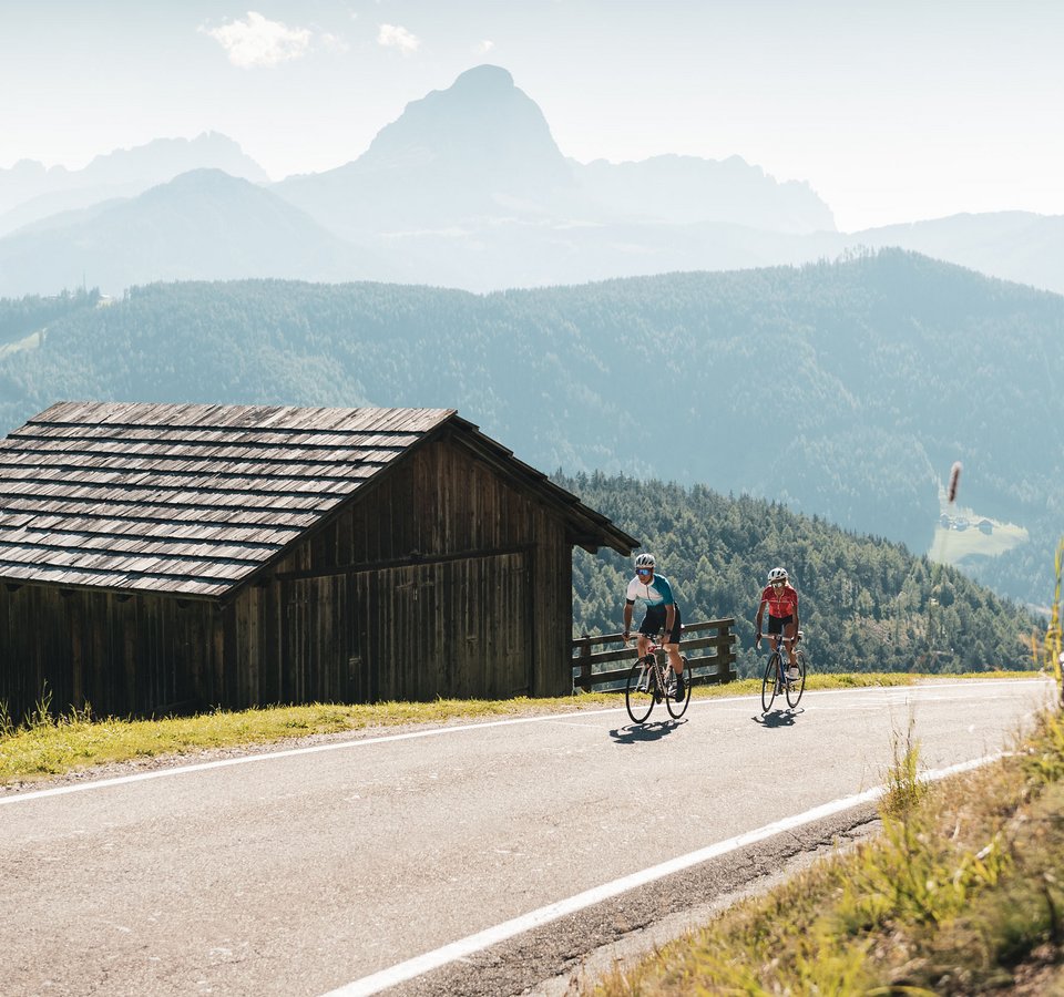 Kronplatz: Höhenmeter ohne Ende © Alex Moling Zwei Radfahrer fahren auf Bergstraße neben Holzhütte in den Alpen