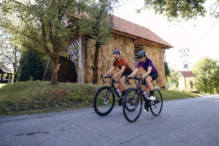 Due ciclisti pedalano su strada di campagna vicino a fienile e alberi