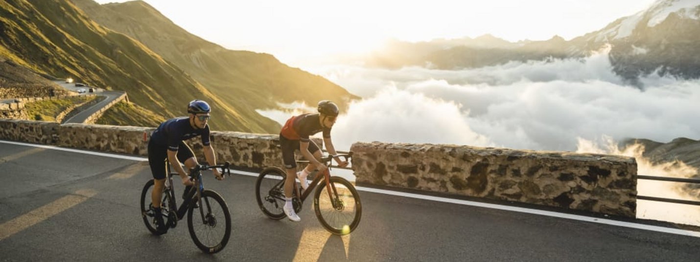 Two cyclists riding on mountain road above clouds at sunrise