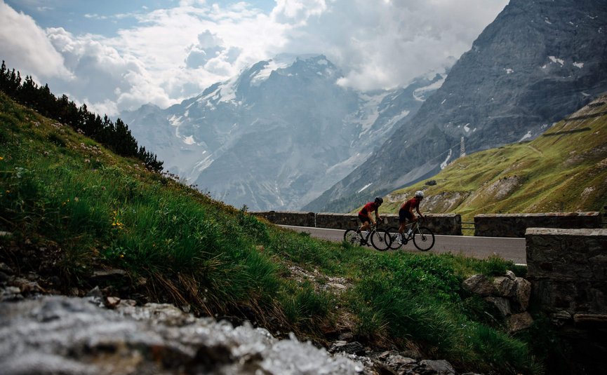 Two cyclists riding on mountain road with cloudy mountains in background