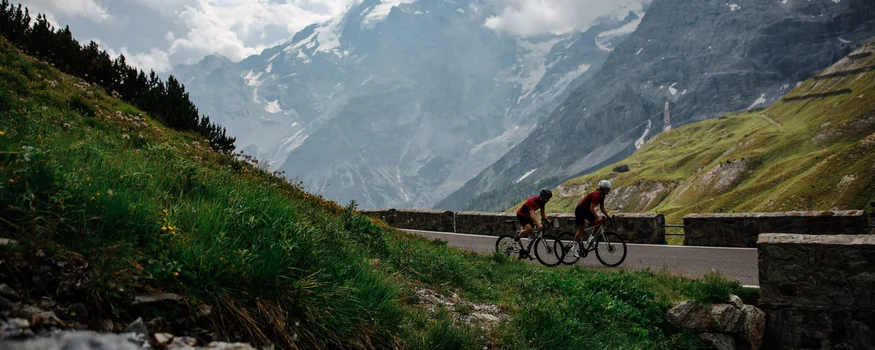 Zwei Radfahrer fahren auf Bergstraße mit bewölkten Bergen im Hintergrund