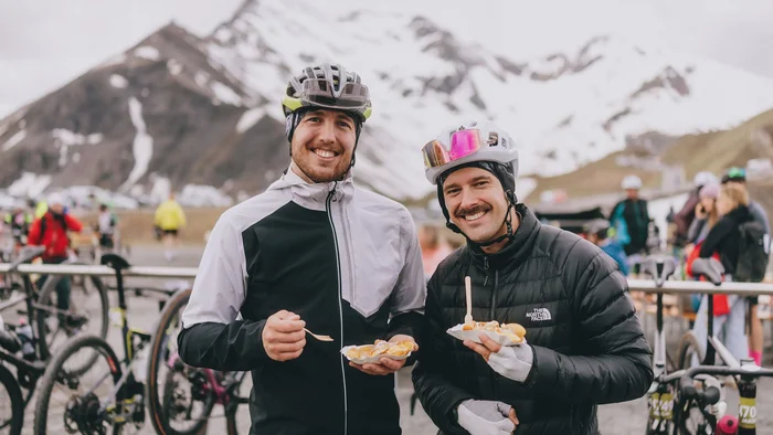 Glocknerkönig 2026 © BRUCK FUSCH GROSSGLOCKNER JFK Photography Zwei Radfahrer mit Helmen essen im Berggebiet vor schneebedeckten Gipfeln
