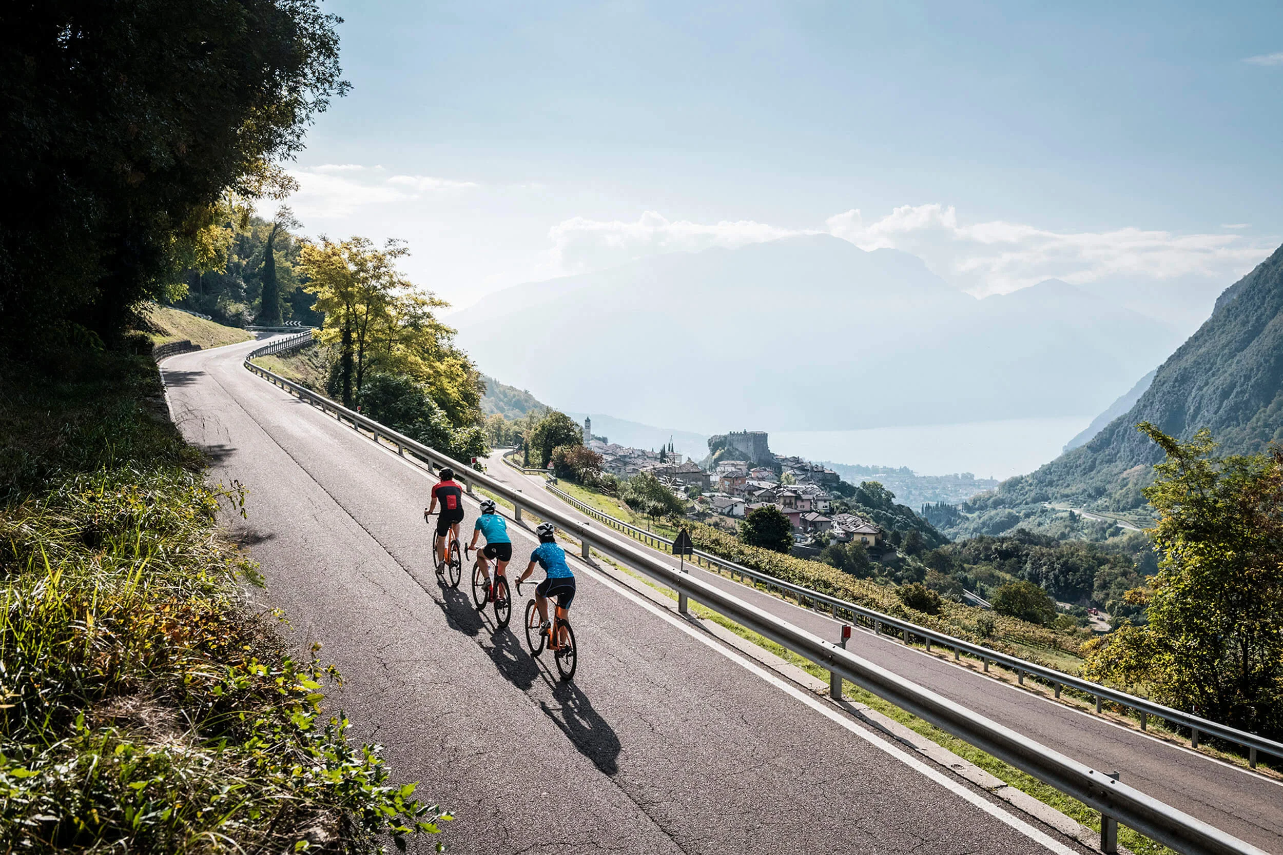 Destinazioni © Alex Moling Tre ciclisti su strada di montagna con vista su paese e lago