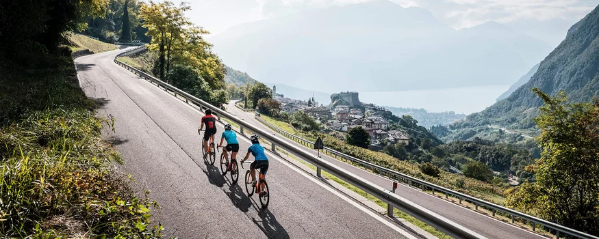 Tre ciclisti su strada di montagna con vista su paese e lago