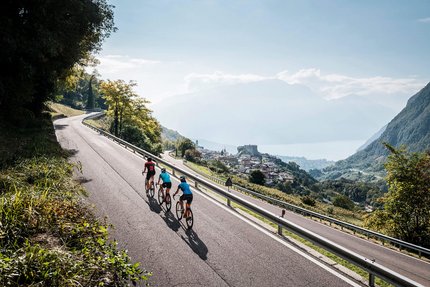 Drei Radfahrer fahren auf Bergstraße mit Blick auf Dorf und See