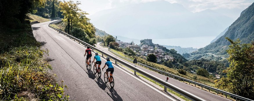 Drie wielrenners rijden op bergweg met uitzicht op dorp en meer
