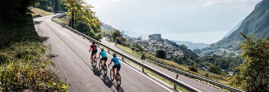 Tre ciclisti su strada di montagna con vista su paese e lago